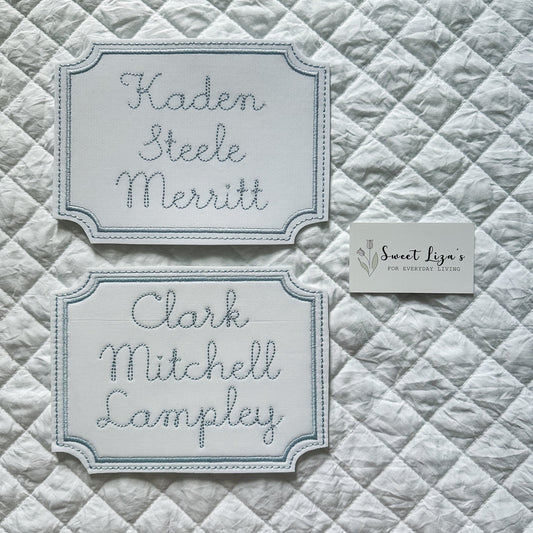 Two embroidered nameplates on a quilted fabric background with 'Sweet Liza's' branding.
